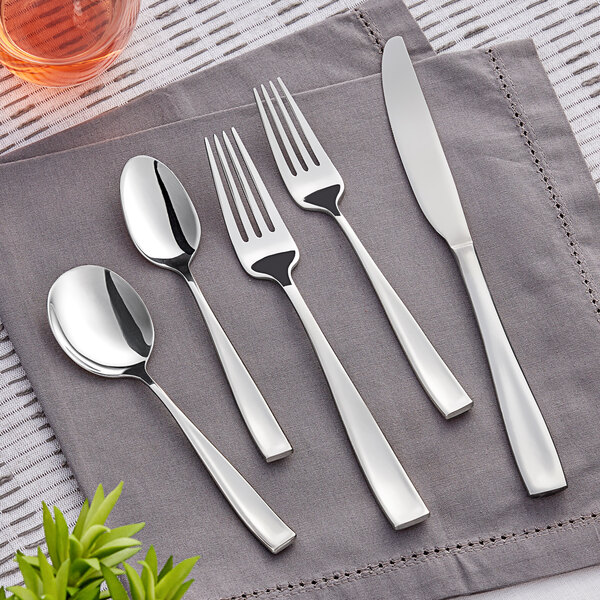 A five-piece set of stainless steel flatware including a knife, two forks, and two spoons, displayed on a gray napkin.