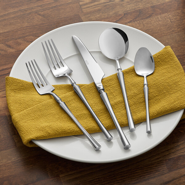 A five-piece set of Acopa Hepburn 18/10 stainless steel flatware, including two forks, a knife, and two spoons, displayed on a white plate with a yellow napkin.
