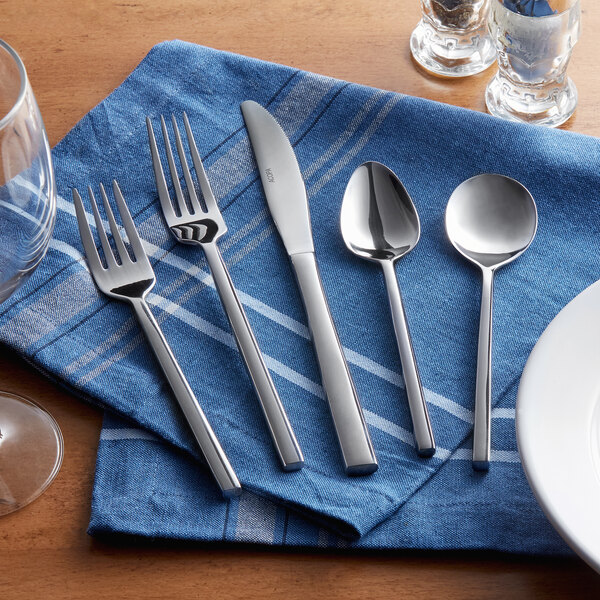 A five-piece set of stainless steel flatware including two forks, a knife, and two spoons, displayed on a blue cloth napkin.