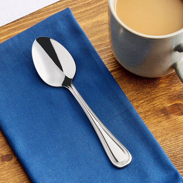 A stainless steel teaspoon with a decorative handle, placed on a blue napkin next to a cup of coffee.