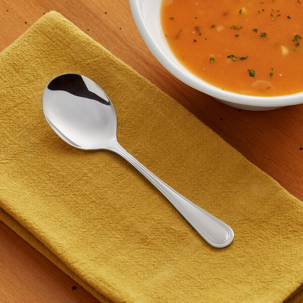 A stainless steel bouillon spoon placed on a yellow napkin next to a bowl of soup.