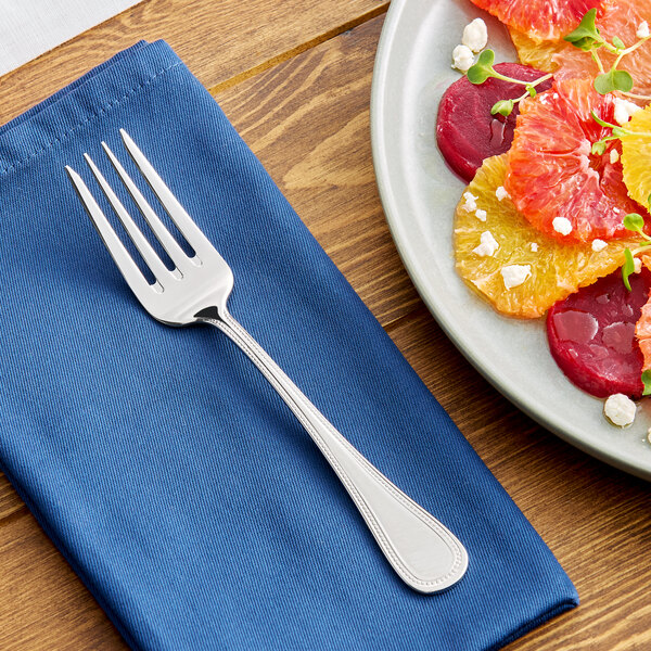 A stainless steel salad or dessert fork placed on a blue napkin next to a plate of food.