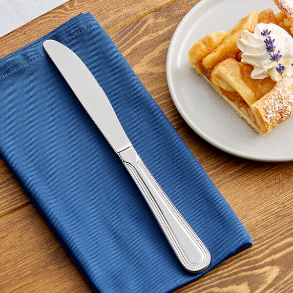 A stainless steel dessert knife placed on a blue napkin next to a plate of dessert.