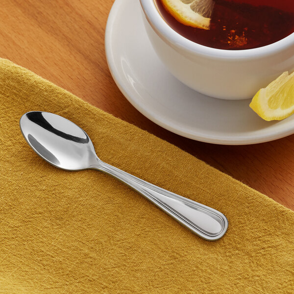 A stainless steel demitasse spoon with a simple, elegant design placed on a yellow napkin next to a cup of tea with lemon.