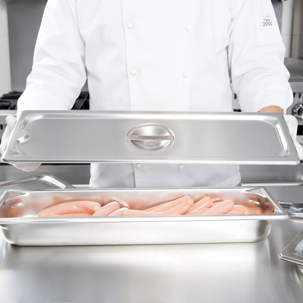 A chef holding a Vollrath stainless steel tray cover over a tray of hot dogs.