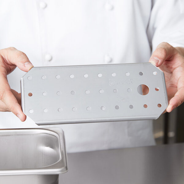 A person holding a Vollrath stainless steel rectangular tray with a metal false bottom with holes.