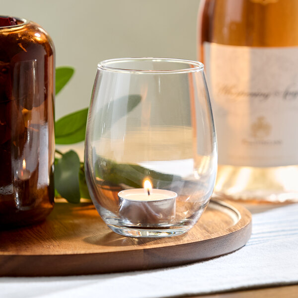 A clear stemless glass with a small lit tealight candle inside, placed on a wooden tray alongside a brown jar and a bottle.