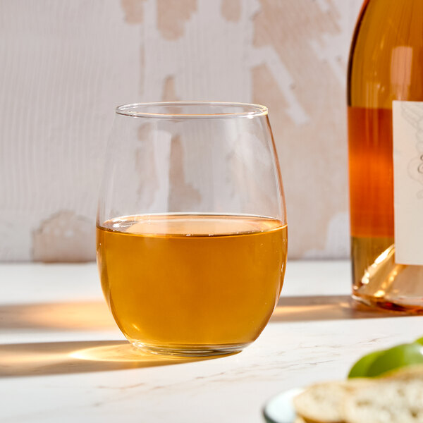 A clear, stemless wine glass filled with a golden liquid, shown on a light surface next to a bottle.