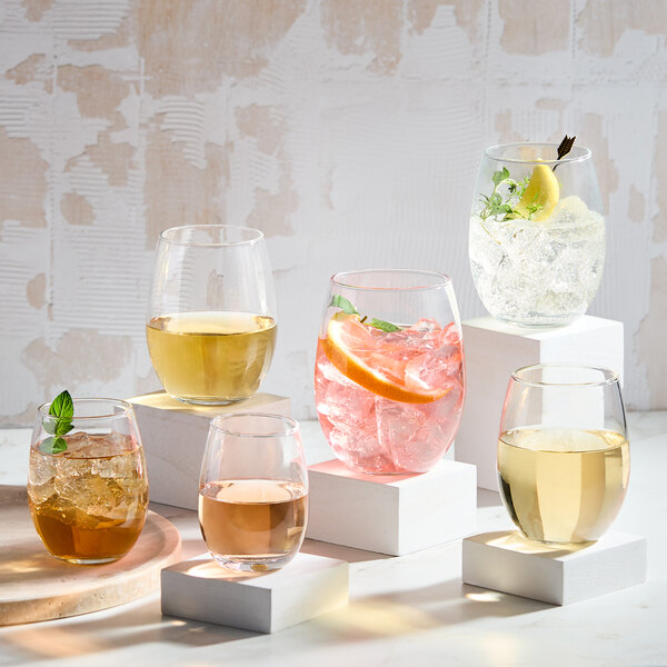 A set of six clear stemless wine tasting glasses, each filled with different beverages and garnishes, displayed on white blocks.
