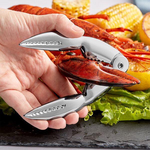A hand is holding a shiny, claw-shaped metal lobster cracker over a cooked lobster and vegetables.