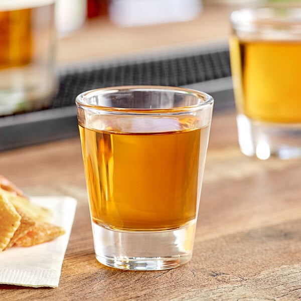 An Acopa shot glass filled with brown liquid next to a plate of crackers.