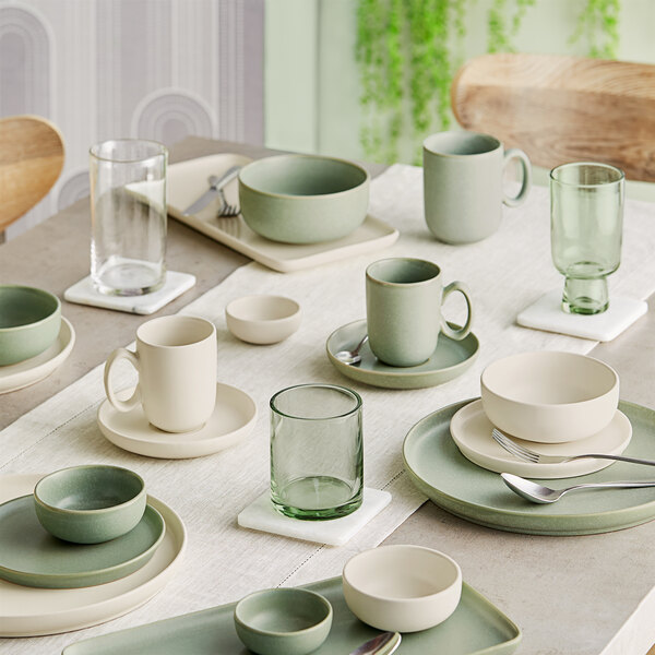 A neatly arranged dining table set with sage green and cream-colored porcelain dishes, bowls, mugs, and green-tinted glassware on a light tablecloth.