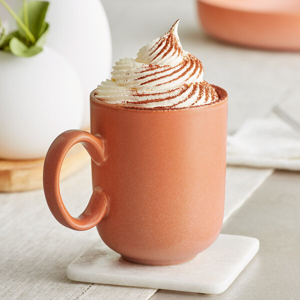 A terra cotta-colored mug filled with a beverage topped with a generous swirl of whipped cream and a dusting of cocoa powder.