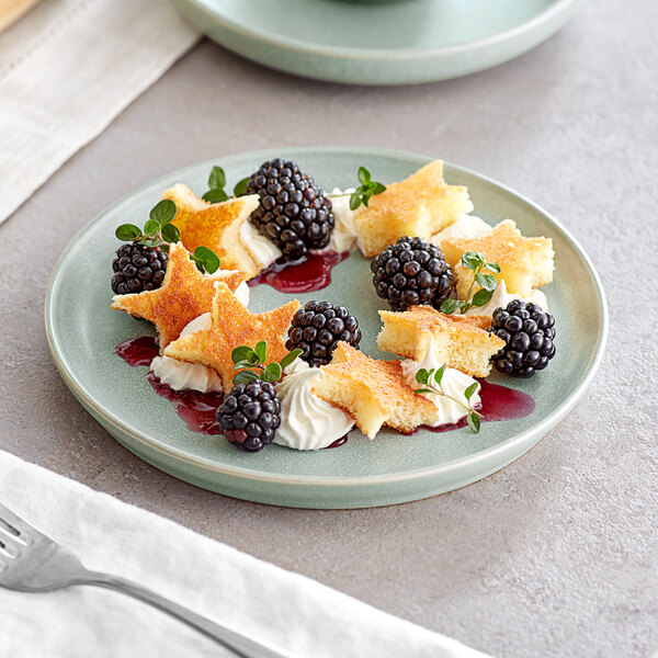A greenish-blue ceramic plate with a dessert arrangement featuring star-shaped cake pieces, blackberries, whipped cream, berry sauce, and small green herbs.
