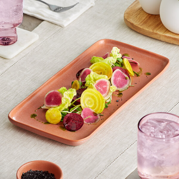 A rectangular terra cotta-colored platter with a matte finish, holding a colorful salad of sliced vegetables and greens.
