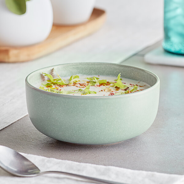 A light blue matte bowl filled with a creamy soup garnished with herbs and spices, placed on a table next to a spoon.