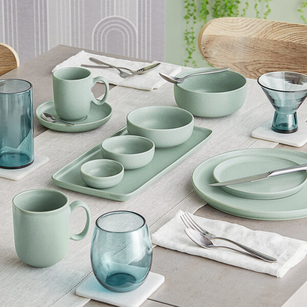A neatly set dining table featuring matte blue-green porcelain dishes, bowls, mugs, and blue-tinted glassware.