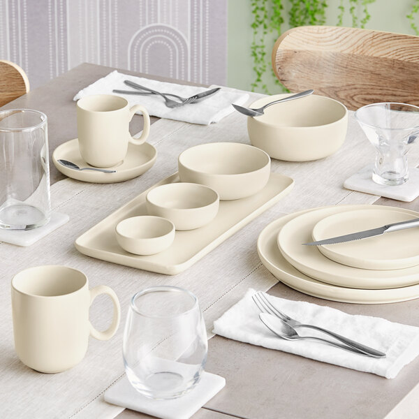 A neatly set dining table featuring cream-colored porcelain dishes, bowls, mugs, and various glassware with silverware placed on white napkins.