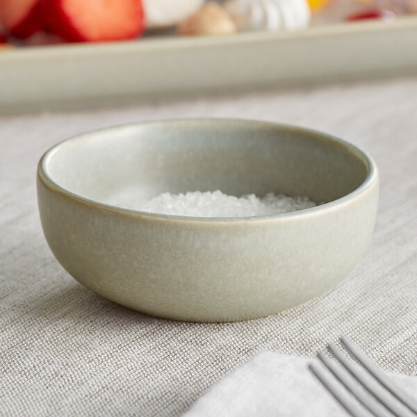 A small, round, ash gray bowl with a matte finish, partially filled with coarse salt, sits on a textured tablecloth.