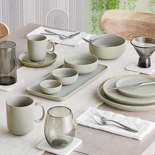 A modern dining table set with ash gray matte finish porcelain dishes, bowls, mugs, and glassware arranged neatly with silverware and white napkins.