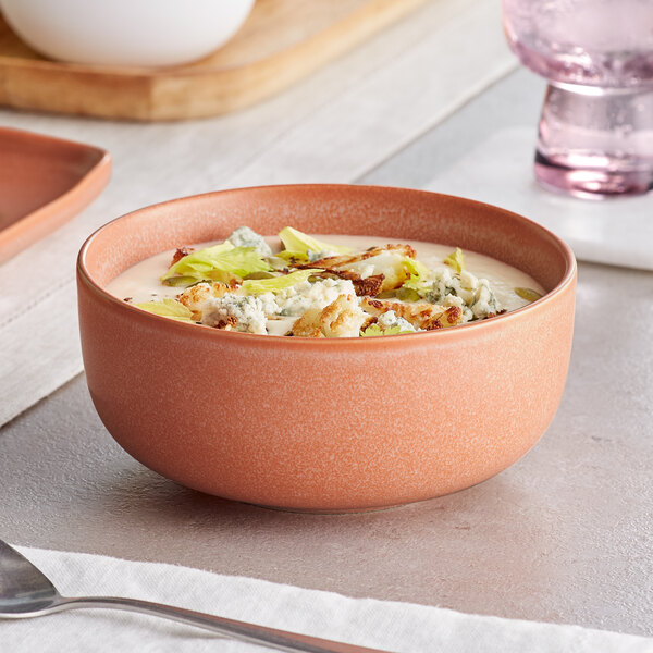 A terra cotta-colored bowl with a matte finish, filled with a creamy soup garnished with greens and crumbled toppings.