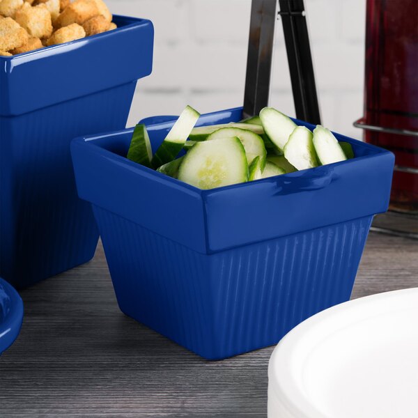 A Tablecraft cobalt blue square container with cucumbers next to a white plate.