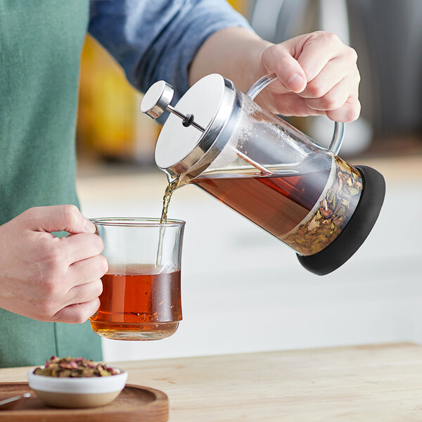 A person is pouring tea or coffee from a glass French press into a clear glass mug.