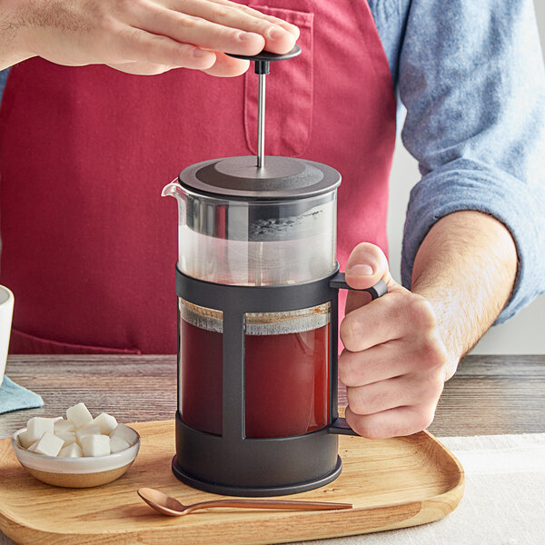 A person is pressing down the plunger of a glass and black French press filled with coffee, with a bowl of sugar cubes and a spoon on a wooden tray nearby.