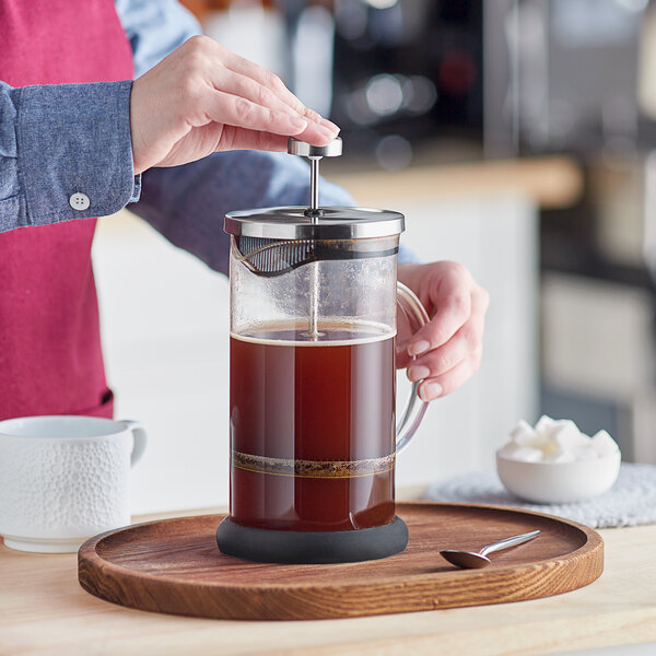 A person is pressing down the plunger of a glass French press filled with coffee on a wooden tray, with a cup, spoon, and bowl of sugar nearby.