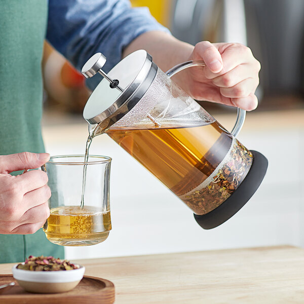 A person is pouring tea or coffee from a glass and metal French press into a clear glass cup.