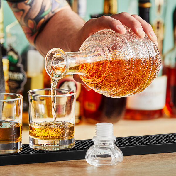 A tattooed person is pouring amber liquid from an ornate, cut-glass decanter into a glass on a bar counter.