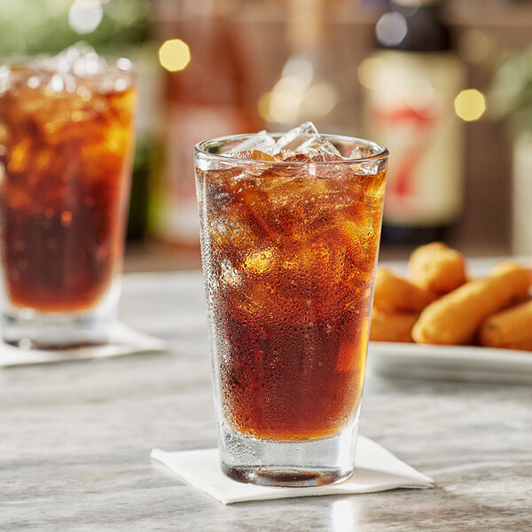 A clear 14 oz. rim tempered mixing glass filled with iced cola, placed on a white napkin.