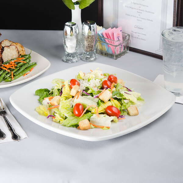 A Tuxton square white china plate with a salad on a table next to a glass of water.