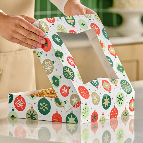 A person is opening a festive bakery box decorated with colorful ornament patterns and a clear window on the lid, revealing a pie inside.