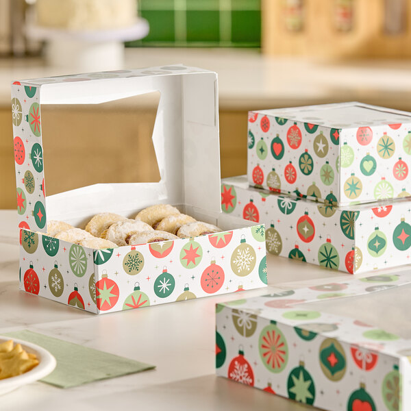 A festive bakery box with a holiday ornament pattern and a clear window on top, partially open to reveal powdered pastries inside, with similar closed boxes stacked nearby.