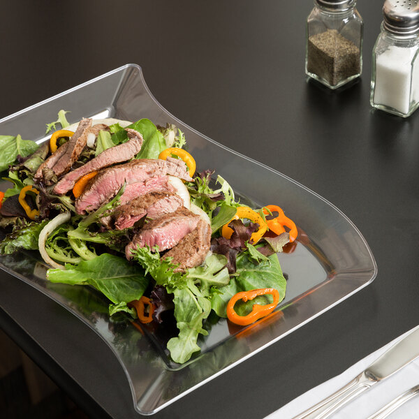 A Fineline clear plastic plate with steak, salad, and vegetables served on it with a knife and fork.