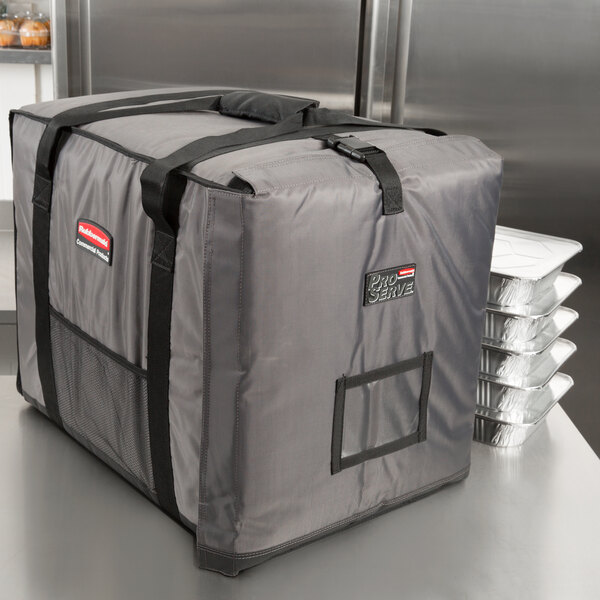 A grey Rubbermaid food pan carrier with black straps sitting next to several trays.