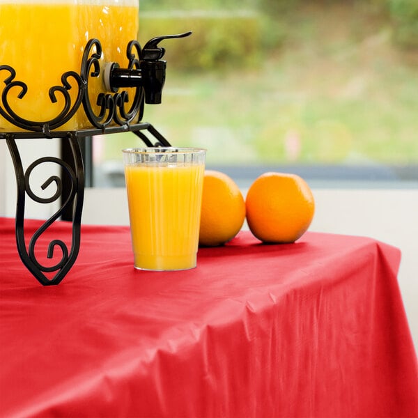 A table with a Creative Converting Classic Red Plastic Tablecover and a drink dispenser with oranges and a glass of orange juice.