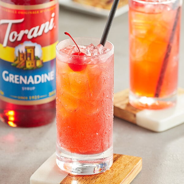 A glass of red liquid with ice and a cherry with a bottle of Torani Grenadine syrup.