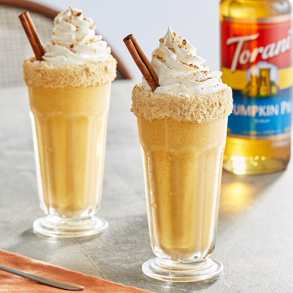 Two glasses of milkshakes with whipped cream and cinnamon sticks made with Torani Pumpkin Pie Syrup on a table.