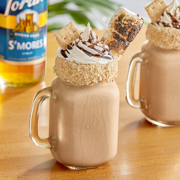 A glass mug of hot chocolate with whipped cream and a cookie on top next to a Torani Sugar-Free S'mores Flavoring Syrup bottle.