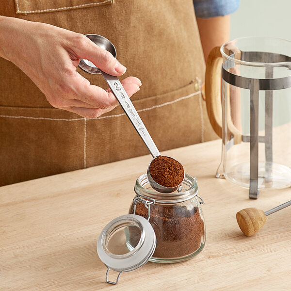 A person using a Tablecraft stainless steel coffee measuring spoon to pour coffee into a jar.