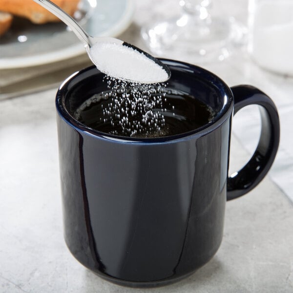 A spoonful of sugar being poured into a cobalt blue CAC Venice stacking mug of liquid.