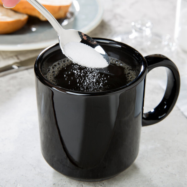 A spoon of sugar being poured into a black CAC Venice mug.