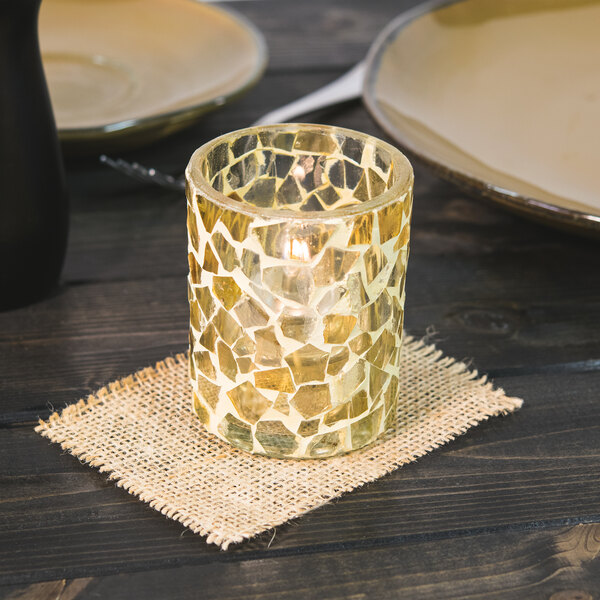 A Sterno light gold mosaic glass votive candle holder on a table with a lit candle inside.