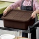 A woman holding a brown Tablecraft bus tub cover over a brown rectangular container on a table.