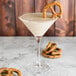 A Chef & Sommelier martini glass filled with a white liquid with a pretzel on the side.