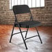 A black National Public Seating metal folding chair with a midnight black padded seat in front of a brick wall.