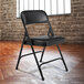 A National Public Seating black metal folding chair with black vinyl padding in front of a brick wall.