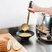 A hand holding a Vollrath stainless steel ladle with a black handle pouring soup into a bowl.